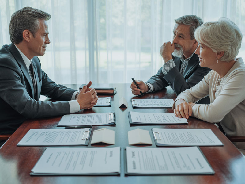 Three people sit at a table with clipboards and documents, engaged in a formal discussion in a well-lit room with large windows.