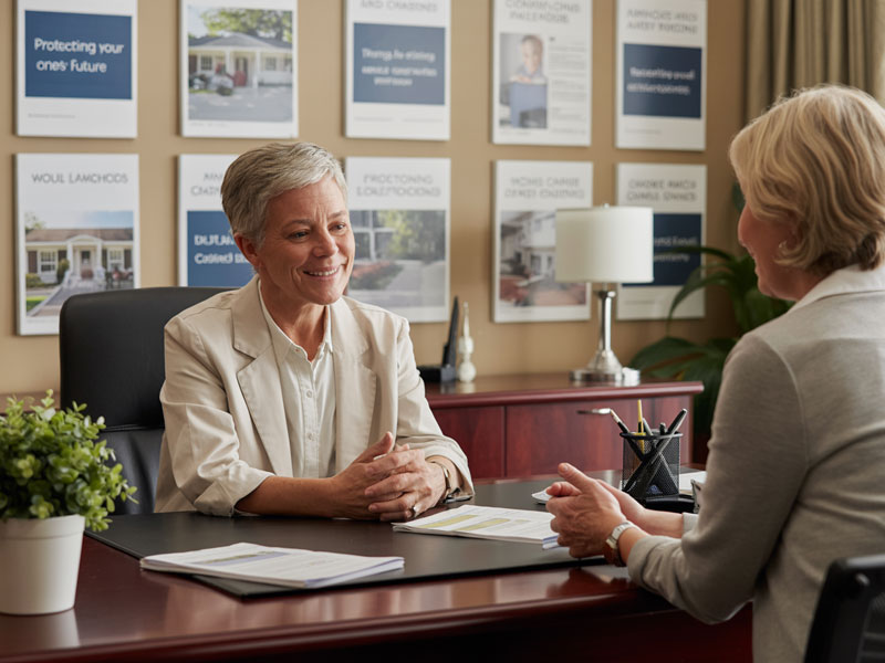 Two women have a discussion at an office desk, with brochures and informational posters displayed on the wall in the background.