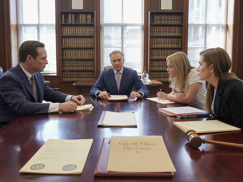 Four people in business attire sit around a conference table with legal documents and a gavel, having a serious discussion in a law library or office.