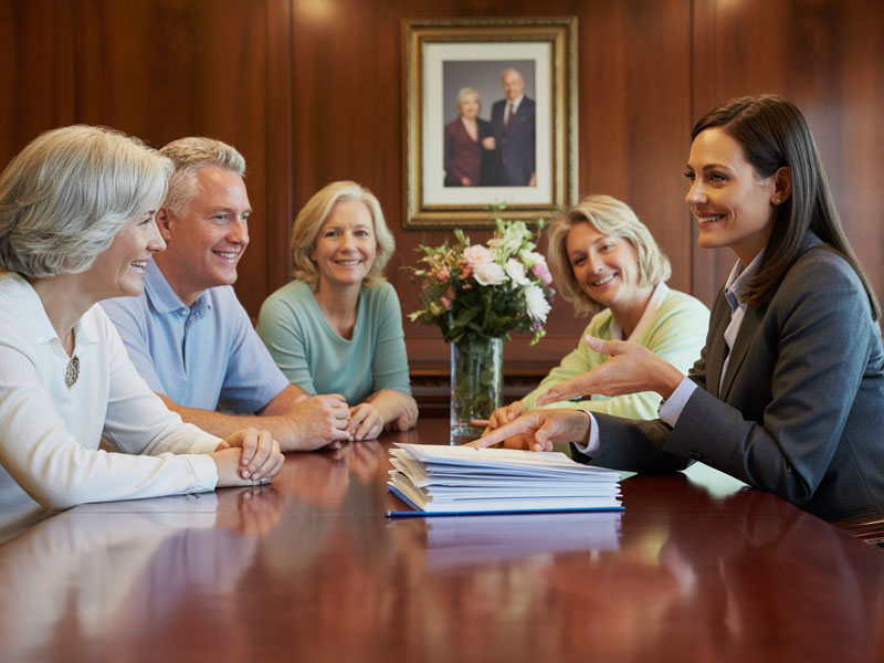 Five adults sit around a conference table with documents, having a discussion in a wood-paneled meeting room with a framed photo and flowers in the background.