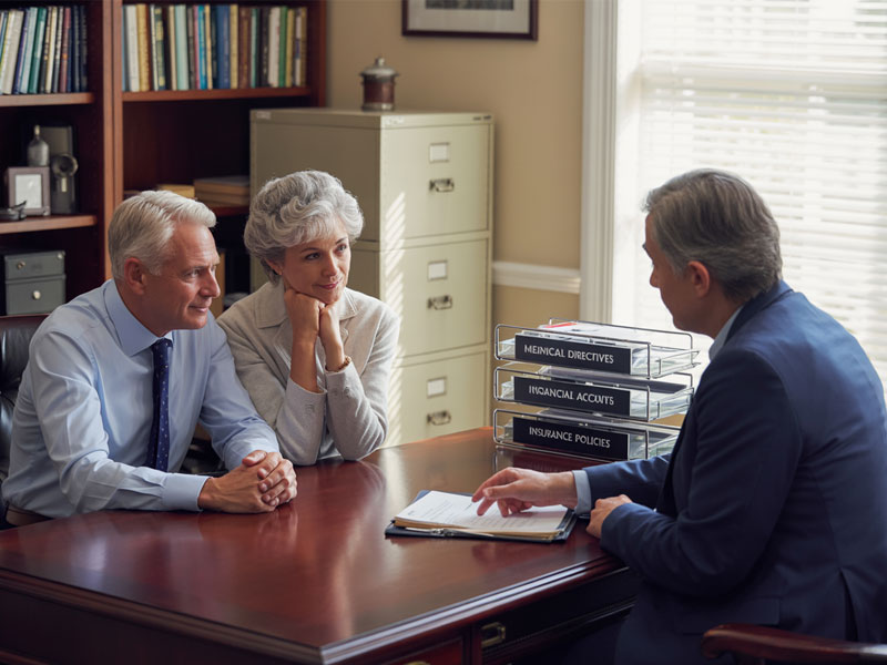 An older couple sits across a desk from a professional who is presenting documents; binders labeled medical directives, financial accounts, and insurance policies are stacked nearby.