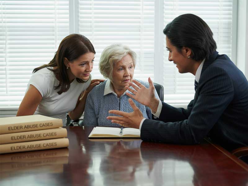 An elderly woman sits between a younger woman and a man in a suit, with law books labeled "Elder Abuse Cases" on the table, suggesting a legal consultation.