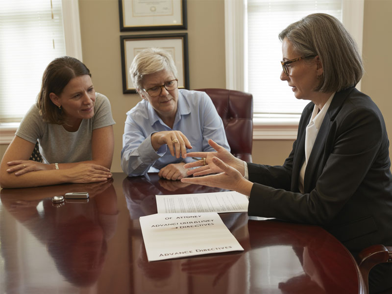 Three women sit at a conference table discussing legal documents labeled "Advance Directives" and "Power of Attorney" in an office setting.