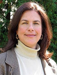 A woman with shoulder-length brown hair wearing a light turtleneck sweater and a checked blazer stands outside in front of greenery.