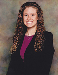 Woman with long curly hair wearing a dark blazer over a purple top, smiling at the camera against a neutral mottled background.